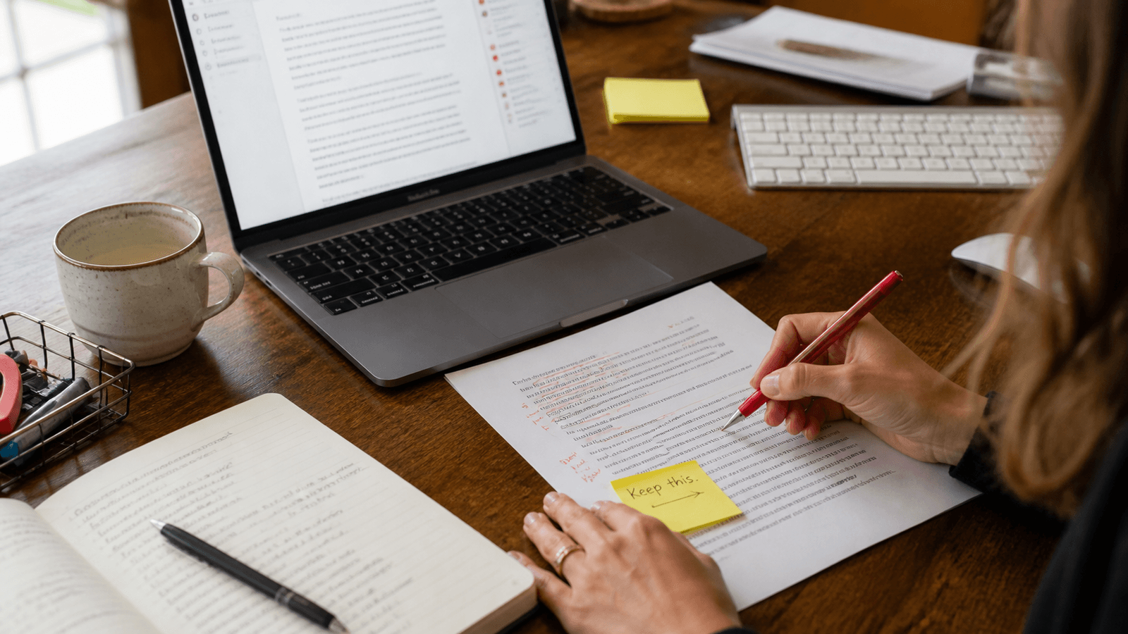 A person marking up a printed draft beside a Mac, with a yellow sticky note reading Keep this on the strongest passage