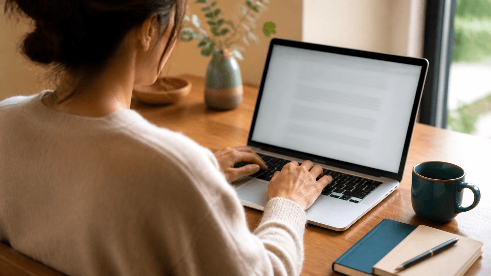 A person types on a laptop at a tidy wooden desk beside a teal mug in warm natural window light.