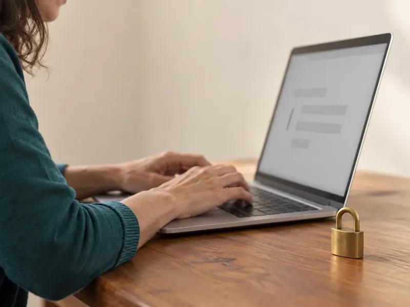 A writer in a teal sweater types on a silver laptop at a wooden desk beside a small brass padlock.