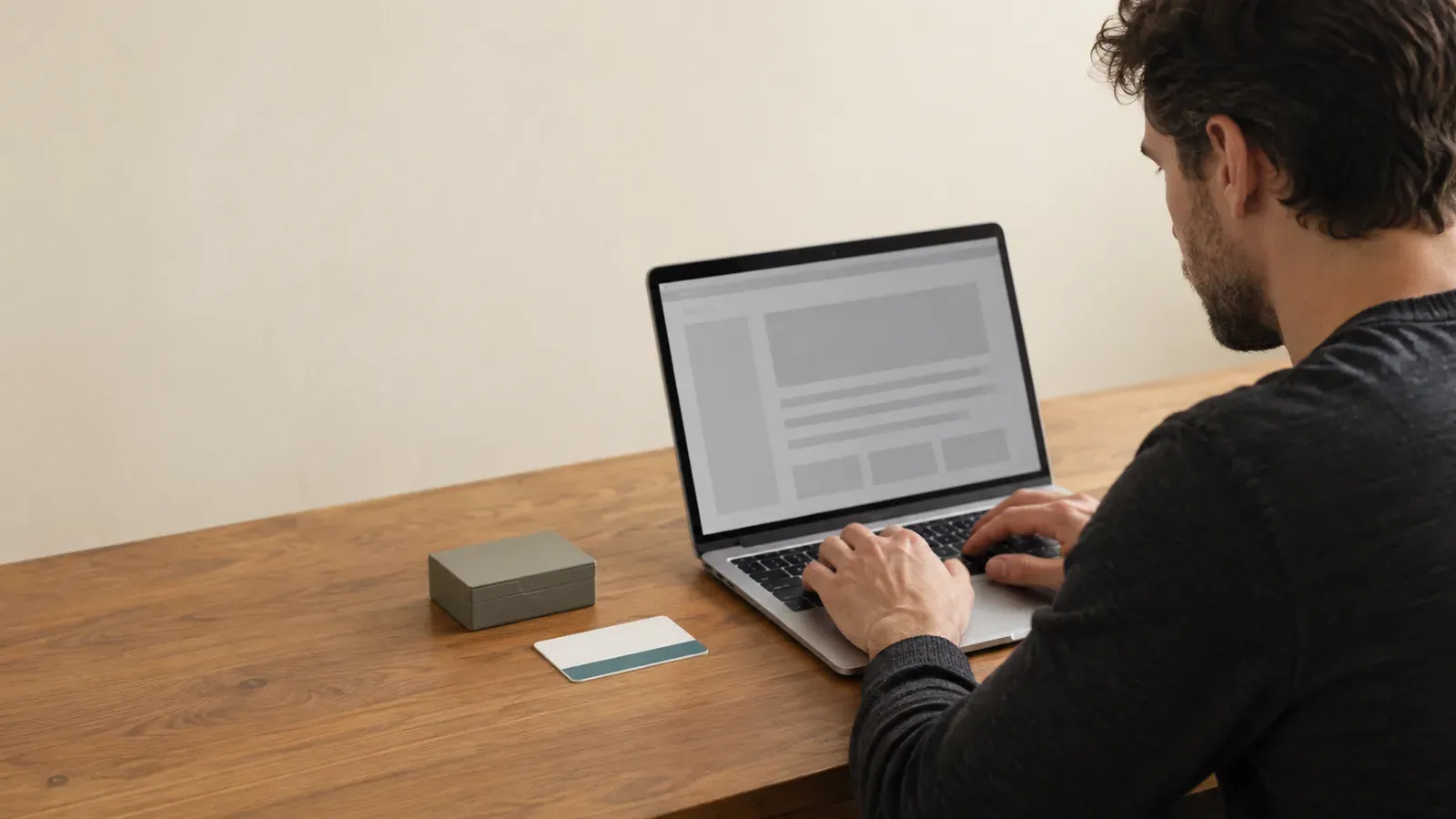 A knowledge worker types on a silver laptop at a wooden desk beside a blank name badge and a small index-card box while gray placeholder blocks fill the screen.